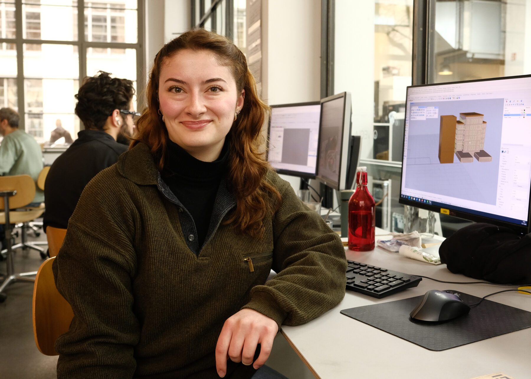 Portrait von Jasmin Dorn - Auszubildende bei werk5. Sie sitzt am Computer hat langes braunes Haar und trägt einen Woll Pullover in grün.