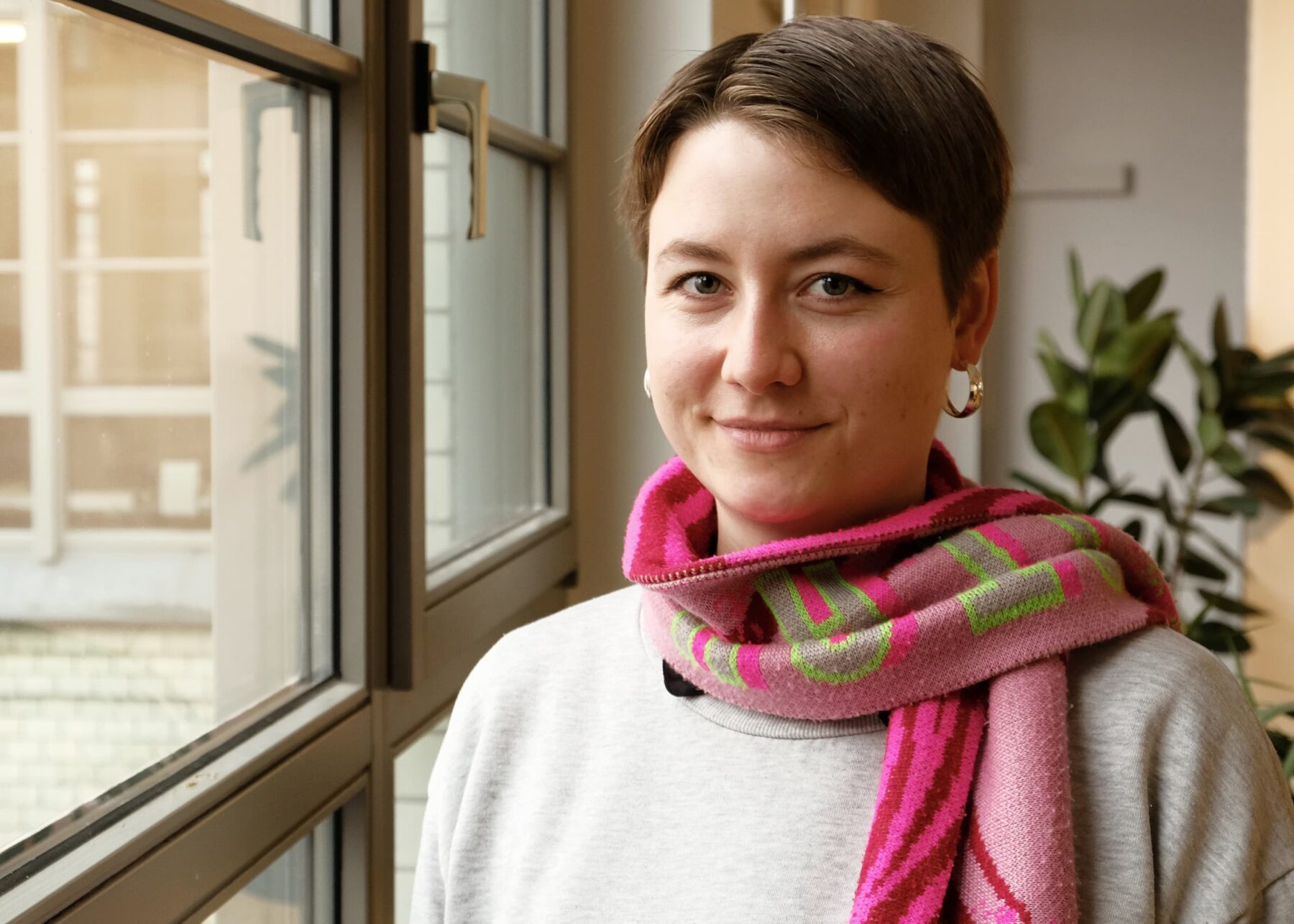 Portrait von leonie Wetter, sie hat kurze braune Haare, grüne Augen und trägt einen Punk/grünen Schal und einen grauen Pullover.